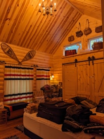 A cozy wooden cabin interior with high wooden ceilings and walls. There is a bed in the foreground with luggage and clothes on it. On the left, there are long striped curtains covering large windows. Snowshoes are mounted on the wall above the windows. A wooden chair and various items are placed beside the windows. The back wall features two small, high windows with baskets on top. There is also a set of barn-style sliding doors in the back.
