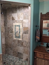 A bathroom featuring a tiled shower area with a decorative tile inlay depicting a bird in the center. The shower tiles are of neutral earth tones. To the right, there's a wood-framed mirror above a wooden cabinet with a towel hanging on a black holder. The room has walls painted in a soft teal color.