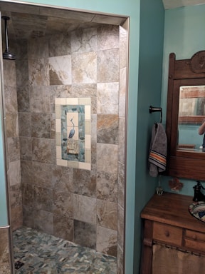A bathroom featuring a tiled shower area with a decorative tile inlay depicting a bird in the center. The shower tiles are of neutral earth tones. To the right, there's a wood-framed mirror above a wooden cabinet with a towel hanging on a black holder. The room has walls painted in a soft teal color.
