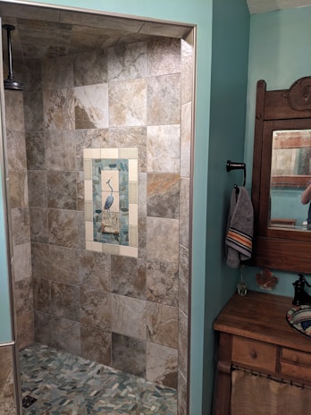 A bathroom featuring a tiled shower area with a decorative tile inlay depicting a bird in the center. The shower tiles are of neutral earth tones. To the right, there's a wood-framed mirror above a wooden cabinet with a towel hanging on a black holder. The room has walls painted in a soft teal color.