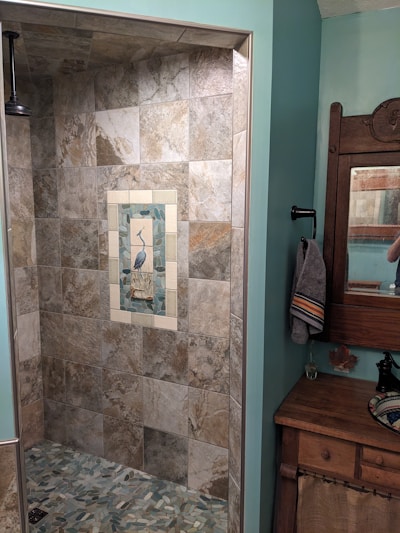 A bathroom featuring a tiled shower area with a decorative tile inlay depicting a bird in the center. The shower tiles are of neutral earth tones. To the right, there's a wood-framed mirror above a wooden cabinet with a towel hanging on a black holder. The room has walls painted in a soft teal color.