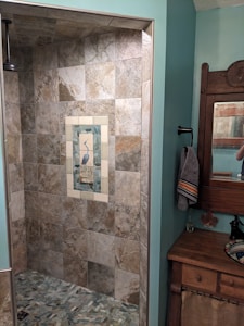 A bathroom featuring a tiled shower area with a decorative tile inlay depicting a bird in the center. The shower tiles are of neutral earth tones. To the right, there's a wood-framed mirror above a wooden cabinet with a towel hanging on a black holder. The room has walls painted in a soft teal color.
