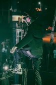 A person in a dark outfit and sunglasses is on stage, energetically singing or speaking into a microphone. The setting is dimly lit with a spotlight on the performer, and a cheering crowd can be seen below.