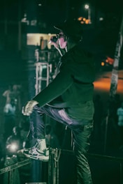 A person in a dark outfit and sunglasses is on stage, energetically singing or speaking into a microphone. The setting is dimly lit with a spotlight on the performer, and a cheering crowd can be seen below.
