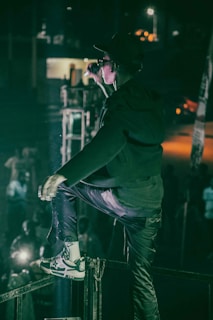 A person in a dark outfit and sunglasses is on stage, energetically singing or speaking into a microphone. The setting is dimly lit with a spotlight on the performer, and a cheering crowd can be seen below.