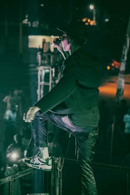 A person in a dark outfit and sunglasses is on stage, energetically singing or speaking into a microphone. The setting is dimly lit with a spotlight on the performer, and a cheering crowd can be seen below.