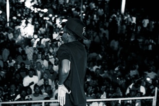 A performer facing a large, densely-packed audience. The main figure is holding a microphone and wearing a cap and sunglasses. The crowd appears engaged and lively, with many subjects slightly blurred, suggesting movement or excitement.