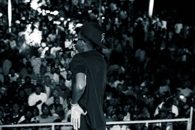A performer facing a large, densely-packed audience. The main figure is holding a microphone and wearing a cap and sunglasses. The crowd appears engaged and lively, with many subjects slightly blurred, suggesting movement or excitement.
