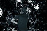 A performer, wearing a cap and sunglasses, is on stage facing a large, densely packed crowd. The scene is in black and white, highlighting the energy and excitement of a live performance with the audience engaging and taking photos.