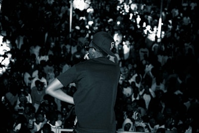 A performer, wearing a cap and sunglasses, is on stage facing a large, densely packed crowd. The scene is in black and white, highlighting the energy and excitement of a live performance with the audience engaging and taking photos.