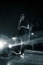 A person stands on a stage holding a microphone, dressed in dark clothing including sunglasses and a cap. Bright stage lights illuminate the scene, creating a spotlight effect and casting shadows. The background is dark, suggesting an event or performance happening at night.