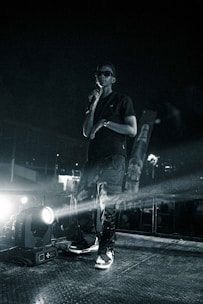 A person stands on a stage holding a microphone, dressed in dark clothing including sunglasses and a cap. Bright stage lights illuminate the scene, creating a spotlight effect and casting shadows. The background is dark, suggesting an event or performance happening at night.
