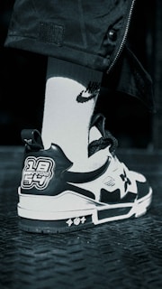 A close-up view of a sneaker with a black and white color scheme featuring unique graphic patches on the back. The shoe rests on a textured dark metal surface, paired with a white sock that has a distinct logo.