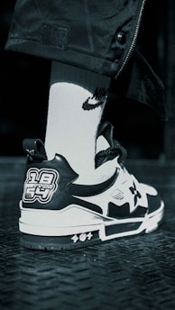A close-up view of a sneaker with a black and white color scheme featuring unique graphic patches on the back. The shoe rests on a textured dark metal surface, paired with a white sock that has a distinct logo.