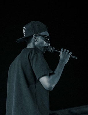 A person is holding a microphone, wearing a cap backwards and sunglasses. The scene is set against a dark background, suggesting a nighttime or indoor concert or performance.