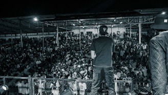 A crowd of people gathered at a nighttime event with a performer on stage facing them. The scene is in black and white, showing a large audience under a covered structure. The performer is standing with their back to the camera, wearing casual clothing, and appears to be engaging with the crowd.