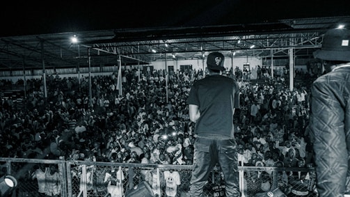 A crowd of people gathered at a nighttime event with a performer on stage facing them. The scene is in black and white, showing a large audience under a covered structure. The performer is standing with their back to the camera, wearing casual clothing, and appears to be engaging with the crowd.