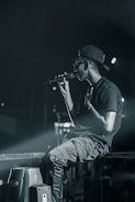 A person wearing a backward cap and sunglasses holds a microphone, appearing to perform on stage. They are seated casually on a metal railing, with a focused and intense demeanor. The background is dimly lit, with spotlights casting a dramatic glow.