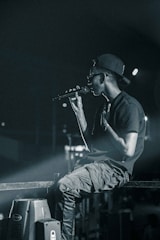 A person wearing a backward cap and sunglasses holds a microphone, appearing to perform on stage. They are seated casually on a metal railing, with a focused and intense demeanor. The background is dimly lit, with spotlights casting a dramatic glow.