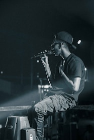 A person wearing a backward cap and sunglasses holds a microphone, appearing to perform on stage. They are seated casually on a metal railing, with a focused and intense demeanor. The background is dimly lit, with spotlights casting a dramatic glow.