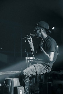 A person wearing a backward cap and sunglasses holds a microphone, appearing to perform on stage. They are seated casually on a metal railing, with a focused and intense demeanor. The background is dimly lit, with spotlights casting a dramatic glow.