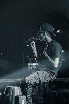 A person wearing a backward cap and sunglasses holds a microphone, appearing to perform on stage. They are seated casually on a metal railing, with a focused and intense demeanor. The background is dimly lit, with spotlights casting a dramatic glow.