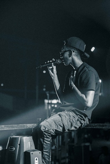 A person wearing a backward cap and sunglasses holds a microphone, appearing to perform on stage. They are seated casually on a metal railing, with a focused and intense demeanor. The background is dimly lit, with spotlights casting a dramatic glow.