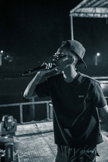A person is on stage holding a microphone, wearing sunglasses and a cap backwards. The setting appears to be a nighttime performance with stage lighting visible in the background.