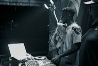 A black and white image featuring a DJ performing at a turntable setup. The DJ raises one hand in the air, signifying engagement with the music. Another person stands nearby, partially visible. Equipment including a laptop and mixer are prominently displayed, illuminated by stage lighting against a dark backdrop.