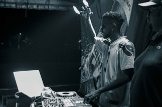 A black and white image featuring a DJ performing at a turntable setup. The DJ raises one hand in the air, signifying engagement with the music. Another person stands nearby, partially visible. Equipment including a laptop and mixer are prominently displayed, illuminated by stage lighting against a dark backdrop.