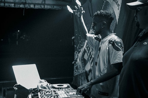 A black and white image featuring a DJ performing at a turntable setup. The DJ raises one hand in the air, signifying engagement with the music. Another person stands nearby, partially visible. Equipment including a laptop and mixer are prominently displayed, illuminated by stage lighting against a dark backdrop.