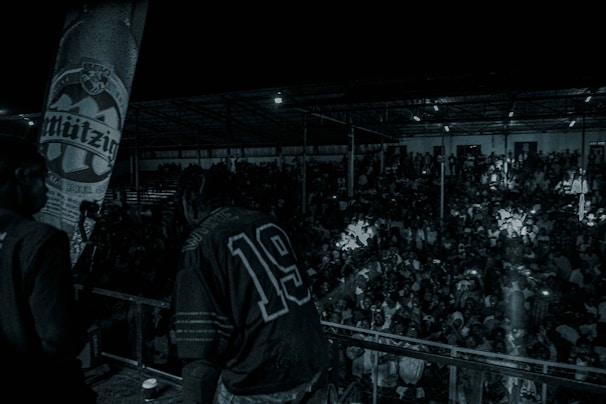 A large crowd gathers in what looks like a concert or event venue. Two individuals are visible in the foreground on a stage, with one wearing a jersey numbered 19. The scene is dimly lit, with only a few bright spots from audience members holding up lights or phones. A large banner is visible to the left.