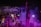 A performer stands on stage facing a large, energetic crowd at a nighttime event. The atmosphere is lively, with many people raising their hands and using their phones to capture the moment. Colored stage lights, predominantly purple, illuminate the scene, and smoke adds to the dynamic ambiance.