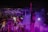 A performer stands on stage facing a large, energetic crowd at a nighttime event. The atmosphere is lively, with many people raising their hands and using their phones to capture the moment. Colored stage lights, predominantly purple, illuminate the scene, and smoke adds to the dynamic ambiance.