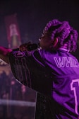 A performer with braided hair holds a microphone on stage, wearing a sports jersey with the name 'WHITE' and the number '1'. The lighting is dim and casts a purplish hue across the scene.
