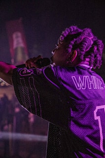 A performer with braided hair holds a microphone on stage, wearing a sports jersey with the name 'WHITE' and the number '1'. The lighting is dim and casts a purplish hue across the scene.