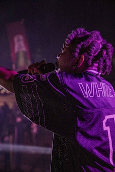 A performer with braided hair holds a microphone on stage, wearing a sports jersey with the name 'WHITE' and the number '1'. The lighting is dim and casts a purplish hue across the scene.