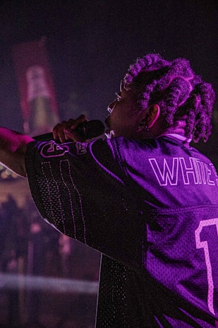 A performer with braided hair holds a microphone on stage, wearing a sports jersey with the name 'WHITE' and the number '1'. The lighting is dim and casts a purplish hue across the scene.