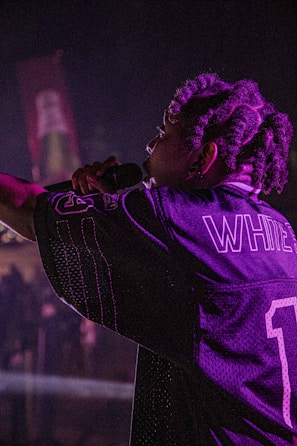 A performer with braided hair holds a microphone on stage, wearing a sports jersey with the name 'WHITE' and the number '1'. The lighting is dim and casts a purplish hue across the scene.