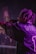 A performer with braided hair holds a microphone on stage, wearing a sports jersey with the name 'WHITE' and the number '1'. The lighting is dim and casts a purplish hue across the scene.