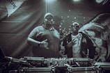 Two individuals stand behind DJ equipment in a dimly lit setting. One person is wearing a polo shirt and cap, while the other is dressed in a jacket and sunglasses. The backdrop features intricate designs and patterns with text elements.