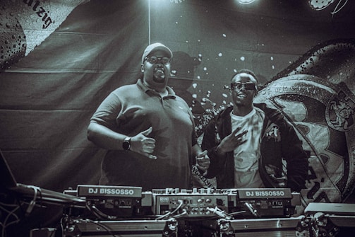 Two individuals stand behind DJ equipment in a dimly lit setting. One person is wearing a polo shirt and cap, while the other is dressed in a jacket and sunglasses. The backdrop features intricate designs and patterns with text elements.