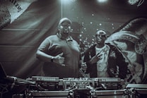 Two individuals stand behind DJ equipment in a dimly lit setting. One person is wearing a polo shirt and cap, while the other is dressed in a jacket and sunglasses. The backdrop features intricate designs and patterns with text elements.