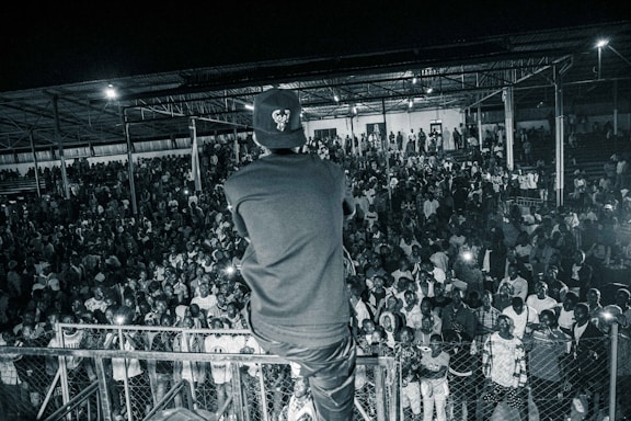 A large crowd is gathered in an indoor venue, with a person standing at the forefront facing the audience. The image is in black and white, and the person appears to be engaging with the crowd from a raised platform. Many people in the audience are looking toward the camera, capturing a lively and dynamic atmosphere.