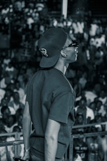 A person wearing a backward cap and sunglasses stands on a stage in front of a large, indistinct crowd. The image has a monochrome filter that creates a moody atmosphere. The person's pose suggests confidence and engagement with the audience.