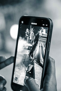 A hand holding a smartphone displaying a black and white image of two people standing near a platform with smoke around them. The phone screen shows a paused video with details such as the time and video quality settings visible. The focus is on capturing the scene through the phone.