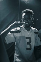 A person wearing sunglasses and an NFL jersey with the number three is in focus. The individual has a short, curly hairstyle and is making a gesture with their right hand. The lighting is low, creating a moody atmosphere with high contrast, and the image is in black and white.