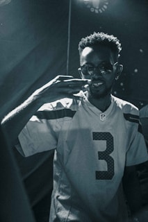 A person wearing sunglasses and an NFL jersey with the number three is in focus. The individual has a short, curly hairstyle and is making a gesture with their right hand. The lighting is low, creating a moody atmosphere with high contrast, and the image is in black and white.