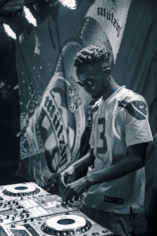 A person wearing a sports jersey operates a DJ mixing console in a dimly lit environment. The background features a banner with abstract designs, and stage lights illuminate part of the scene, creating a focused and moody ambiance.