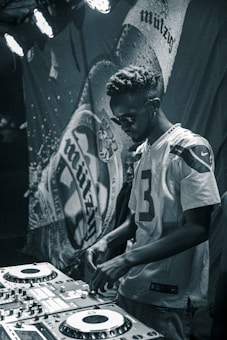 A person wearing a sports jersey operates a DJ mixing console in a dimly lit environment. The background features a banner with abstract designs, and stage lights illuminate part of the scene, creating a focused and moody ambiance.